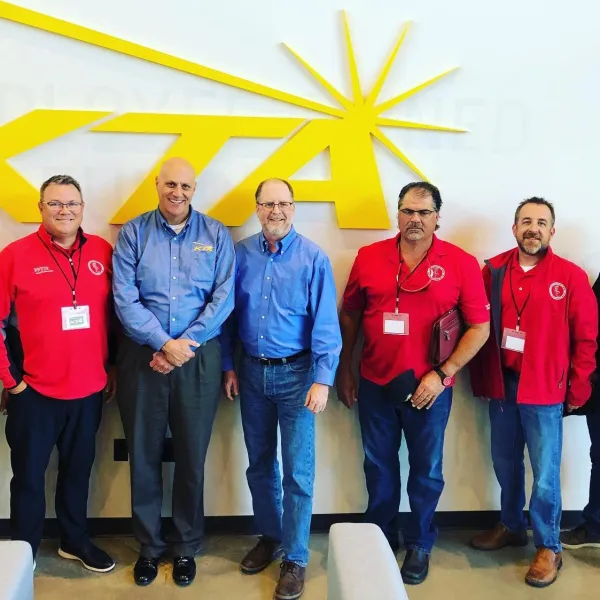 Seven men standing in front of a large yellow KTTA logo on a white wall, dressed in red and blue shirts.