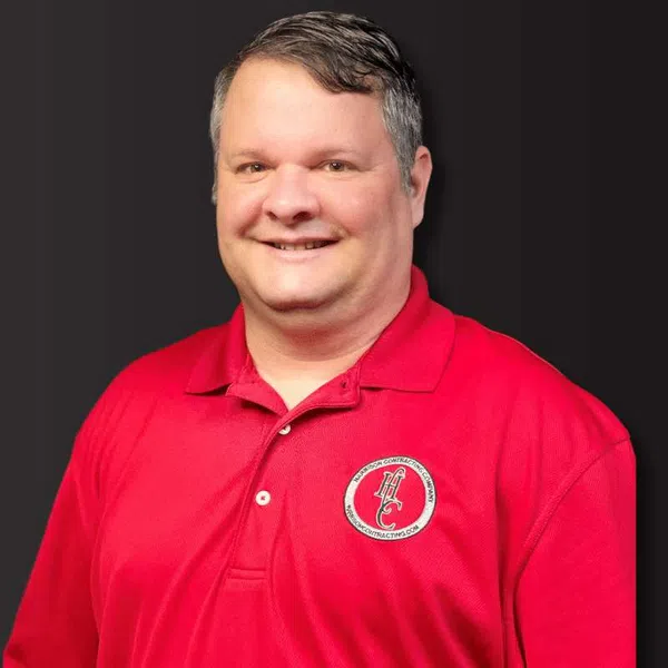 Smiling man wearing a red polo shirt with a circular logo on a dark background.