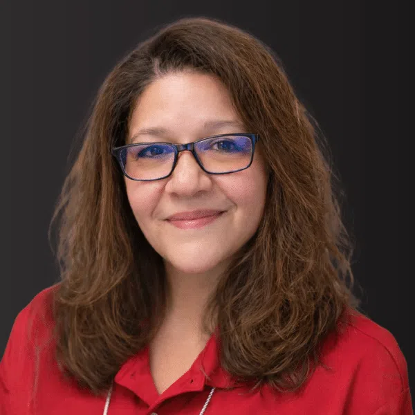 Smiling woman with shoulder-length brown hair wearing glasses and a red collared shirt against a dark background.