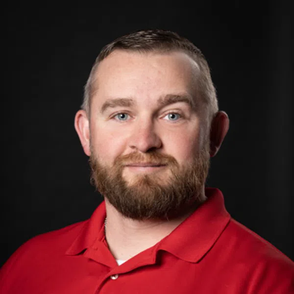 Bearded man wearing a red polo shirt with a round emblem on a black background