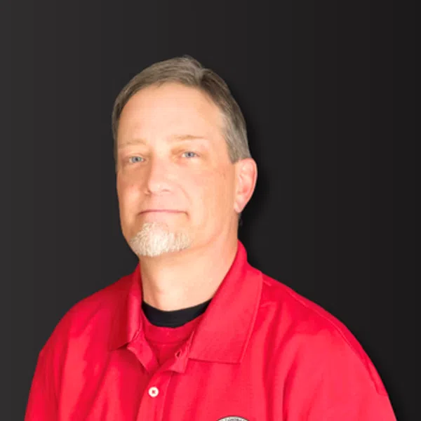 Man with short gray beard wearing a red polo shirt with an embroidered logo on a dark background.