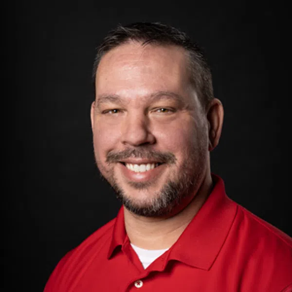 Smiling man with short hair and beard wearing a red polo shirt with an embroidered logo on black background