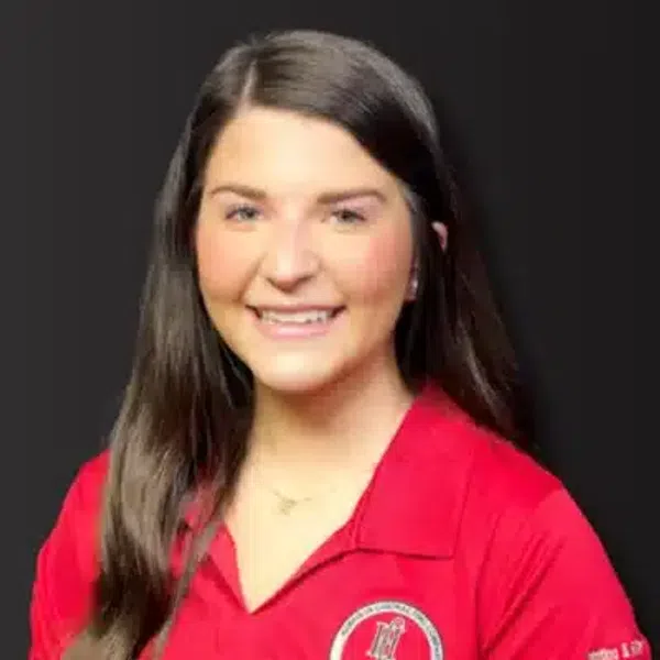Smiling young woman with long brown hair wearing a red polo shirt with a circular logo on chest and sleeve.