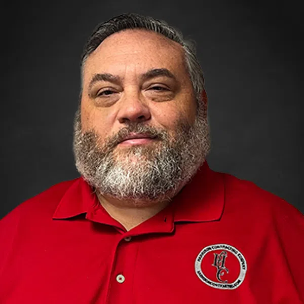 Middle-aged man with gray beard wearing a red polo shirt with a circular embroidered logo over black background