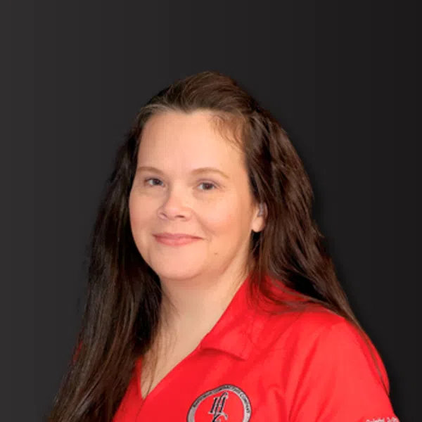 Smiling woman with long brown hair wearing a red polo shirt with an embroidered logo and text on dark background