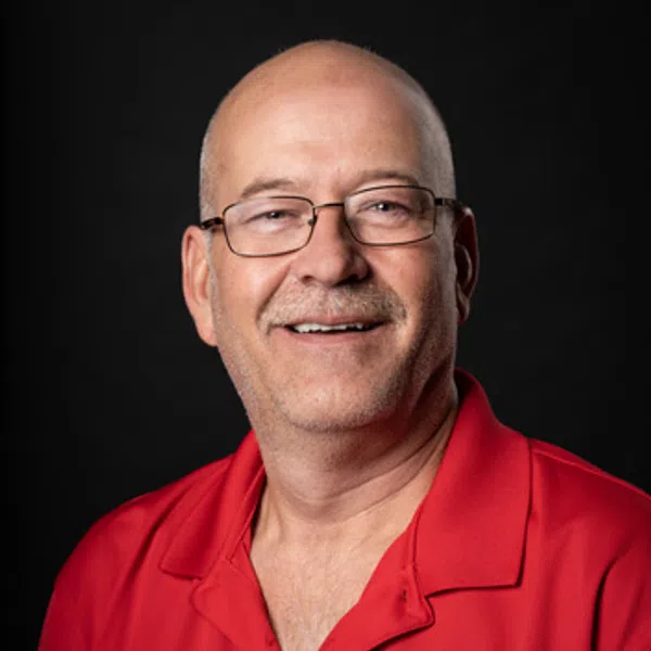 Smiling bald man with glasses wearing a red shirt with a round logo on the chest, black background.