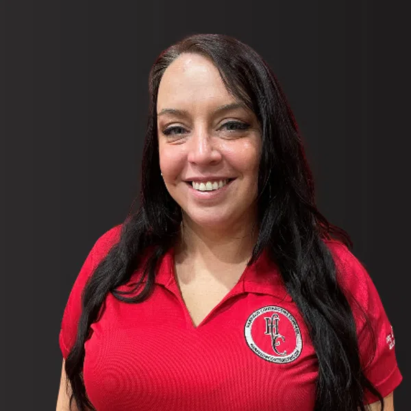 Smiling woman with long dark hair wearing a red polo shirt with a circular logo on the chest against dark background