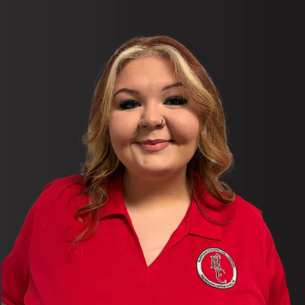 Smiling woman with blond hair wearing a red polo shirt with a circular HC logo on a dark background