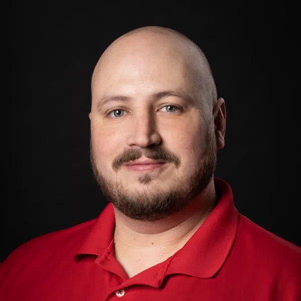 Portrait of a bald man with a beard wearing a red polo shirt with a circular logo on a black background