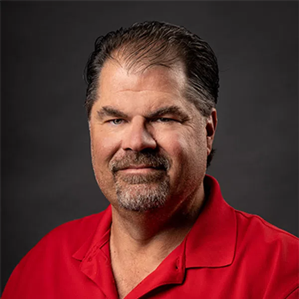 Middle-aged man with short hair and goatee wearing red polo with a circular company logo on black background