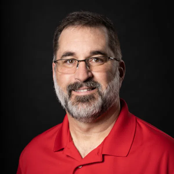 Middle-aged man with glasses and beard wearing red polo with company logo on black background
