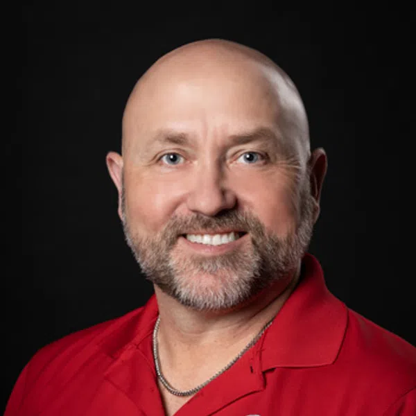 Smiling bald man with a beard wearing a red polo shirt with a logo on a black background.