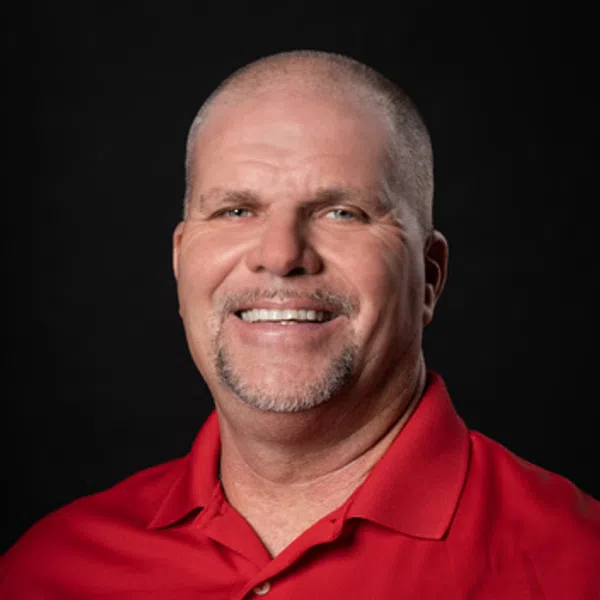 Smiling bald man with goatee wears a red polo shirt with a circular company logo on black background.