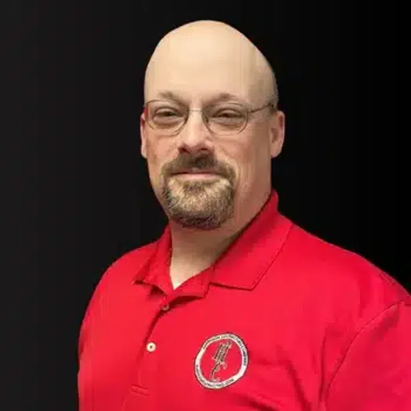 Man with glasses and goatee wearing a red polo shirt with a logo against a black background