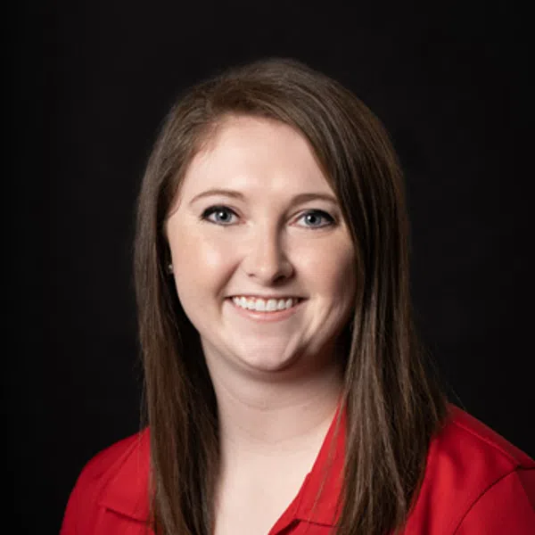 Smiling woman with straight brown hair wearing a red collared shirt against a black background.