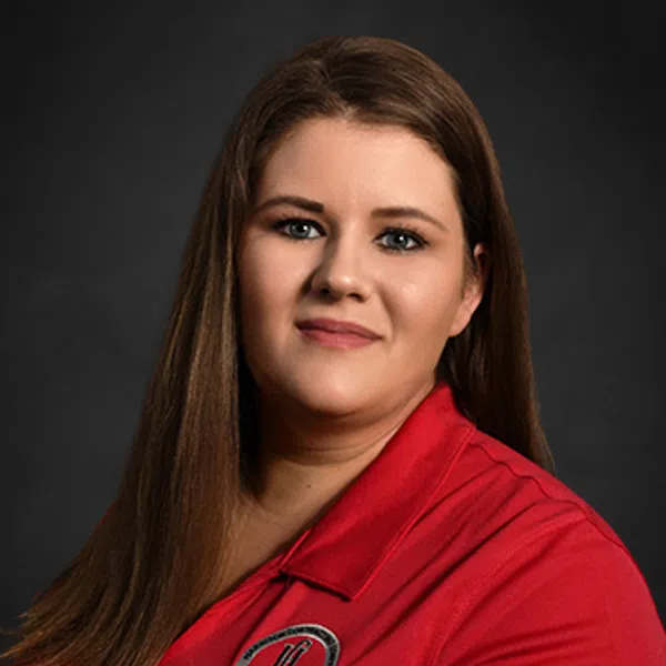 Woman with long brown hair wearing a red polo shirt with embroidery against a dark background.