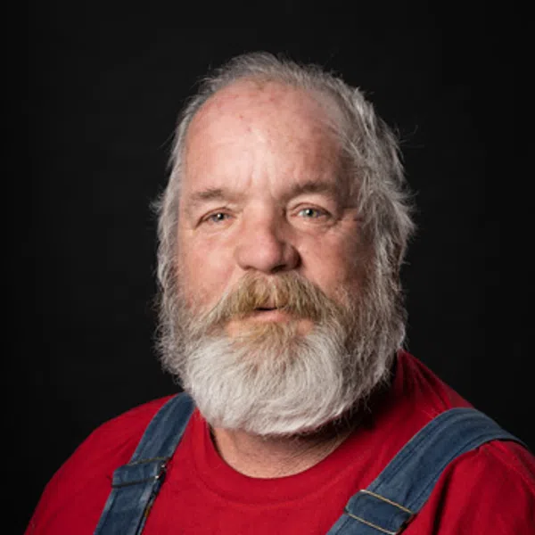 Portrait of a bearded man with gray hair wearing a red shirt and blue denim overalls against a black background