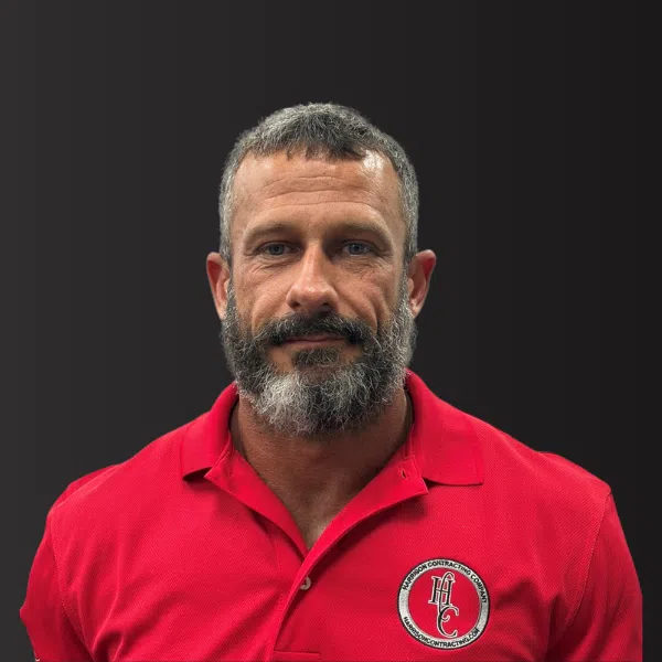 Man with gray beard wearing a red polo shirt with a contractor company logo on a dark background.