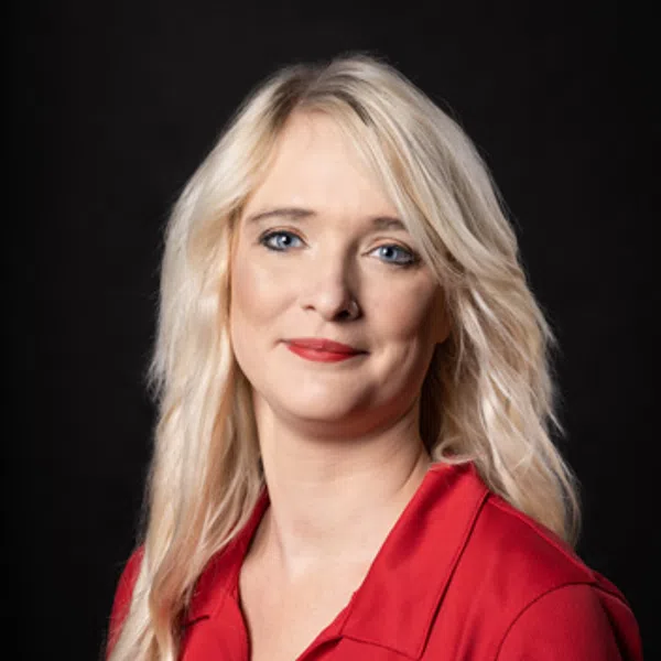 Blonde woman with blue eyes wearing a red collared shirt with an embroidered logo on black background