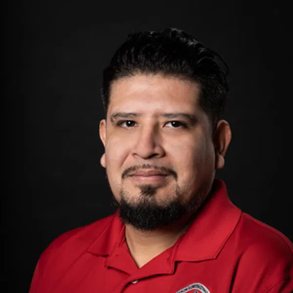 Man with dark hair and beard wearing a red collared shirt with a logo on black background.