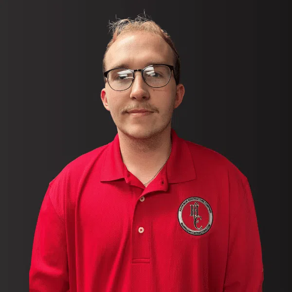 Young man with glasses wearing a red polo shirt with a logo against a dark background.