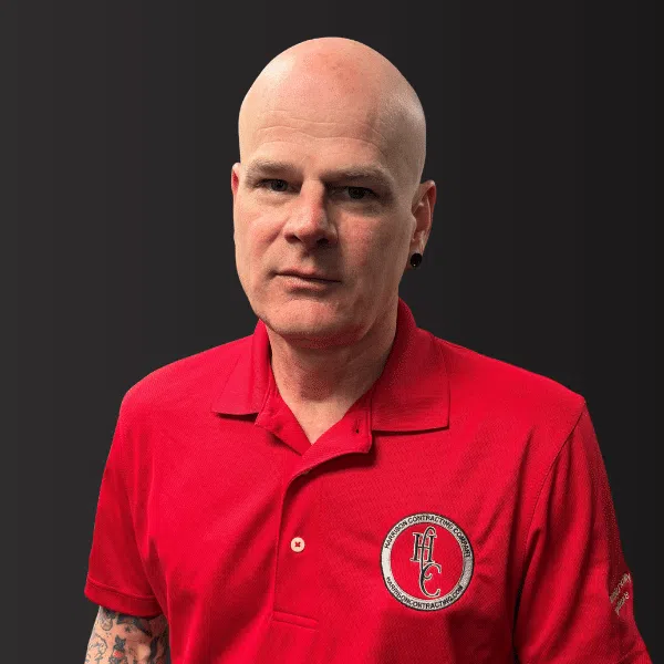 Bald man with tattooed arm wearing a red polo shirt with construction company logo on a dark background