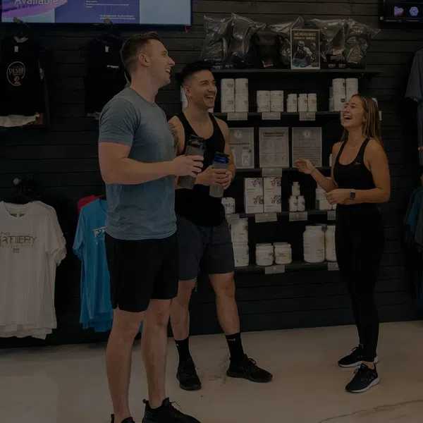 Three young adults in athletic wear smiling and chatting inside a sports nutrition store with supplement jars and apparel.
