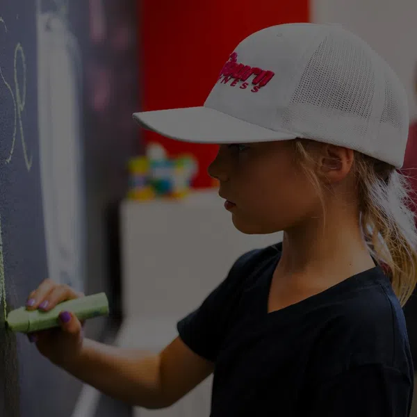 Young girl wearing a white cap writing 'go go' in yellow chalk on a blackboard indoors.