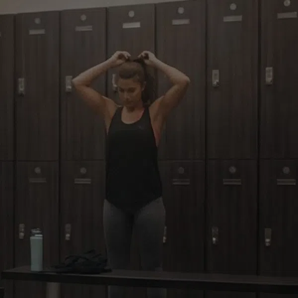 Woman tying hair in a gym locker room with dark lockers and a bench in the foreground.