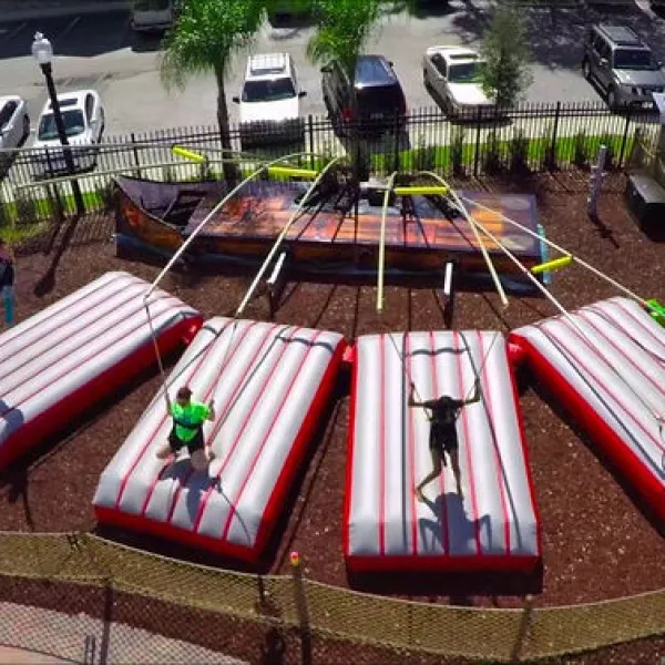 Aerial view of three people bouncing on large inflatable mats connected to bungee cords in an outdoor park.