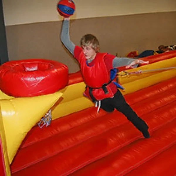 Young person in safety harness playing inflatable basketball game indoor, reaching to score with ball.