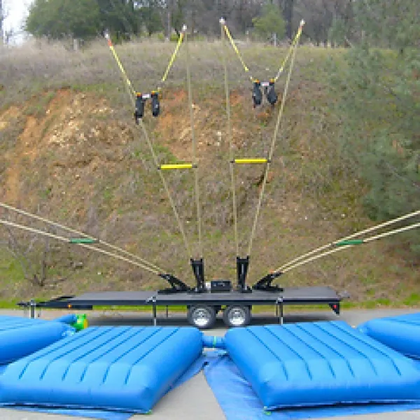 Bungee trampoline setup with blue air cushions and a trailer in an outdoor park area with trees and dirt hillside.