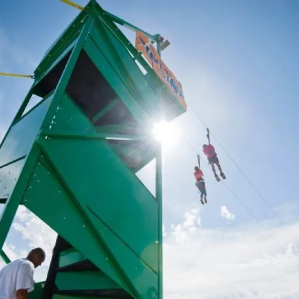 Two people ziplining from a tall green platform under a bright blue sky with scattered clouds.