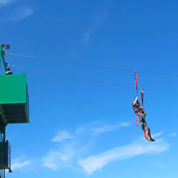 Person wearing safety gear rides a zipline against a bright blue sky near a green platform.