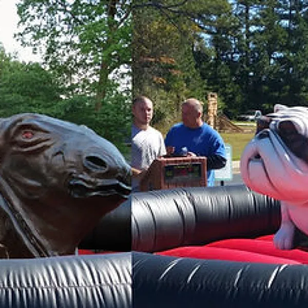 Two women riding mechanical bull and bulldog outdoors at a sunny event with spectators and trees