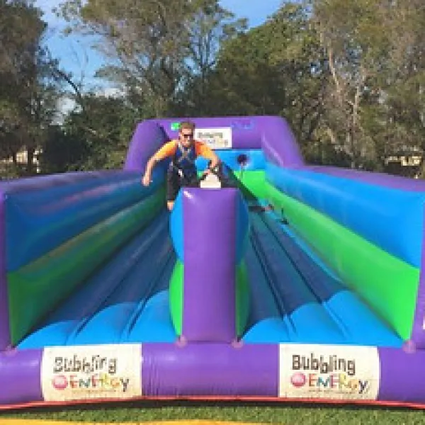 Person balancing on a blue and green inflatable obstacle course with purple sides outdoors on grass.