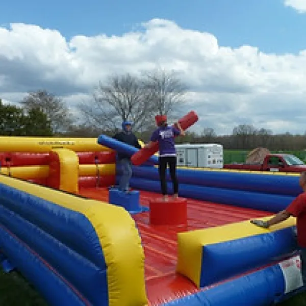 Two people play on a colorful inflatable jousting arena outdoors with a referee watching nearby under a blue sky.