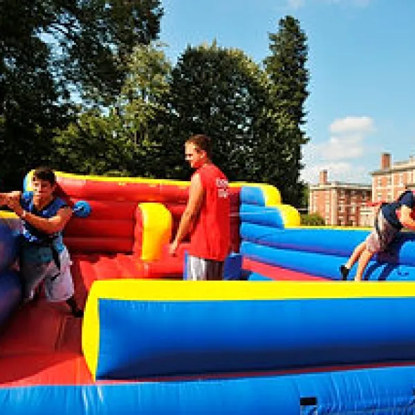 Three people playing on a colorful inflatable obstacle course outdoors with blue sky and trees in background
