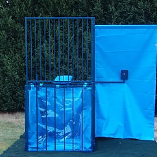 Blue dunk tank with metal cage and splash panel set outdoors on grass near green hedge.