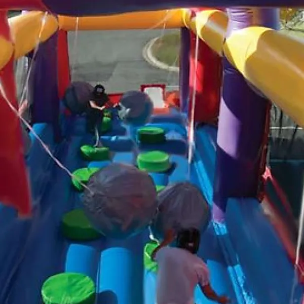 Children playing and dodging large swinging balls on a colorful inflatable obstacle course outdoors.