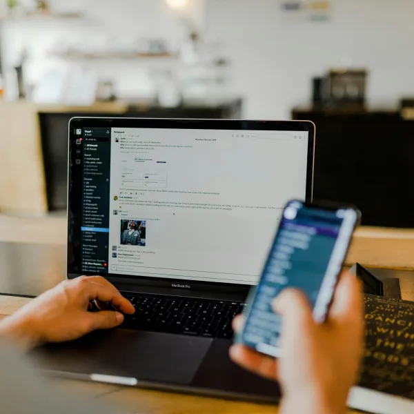 Person using a MacBook laptop and holding a smartphone inside a cozy cafe with blurred background.