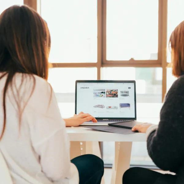 Two women browsing product images on a laptop at a white table in a bright room with large windows.