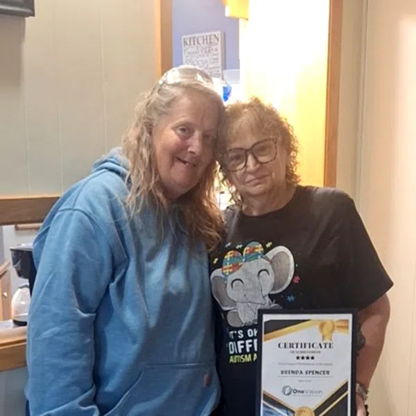 Two women standing close, one holding a framed certificate of recognition indoors in casual clothing.
