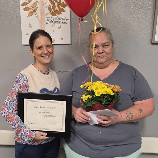 Two women stand together, one holding a framed award and the other holding flowers with balloons in the background.
