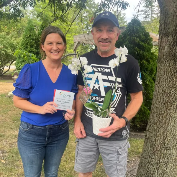 Smiling woman and man outdoors holding an award plaque and a white orchid plant under a tree.