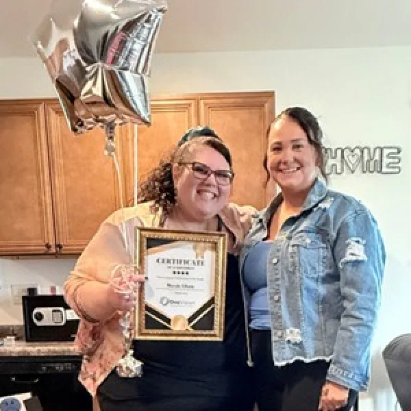 Two smiling women pose indoors, one holding a framed certificate and silver star-shaped balloons, celebrating achievement.