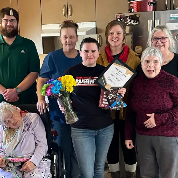 Group of seven people in kitchen, one holding flowers, certificate, and chocolate, celebrating achievement.