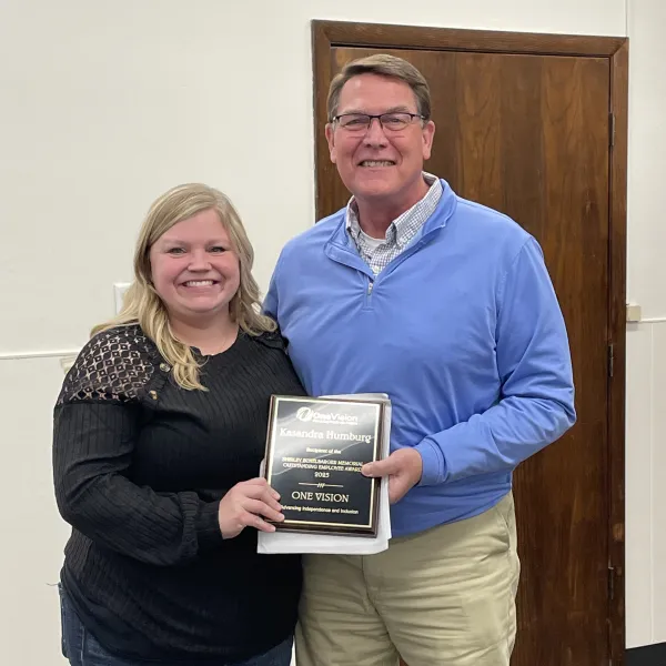 Two people smiling indoors, holding an award plaque recognizing outstanding employee achievement in 2023.
