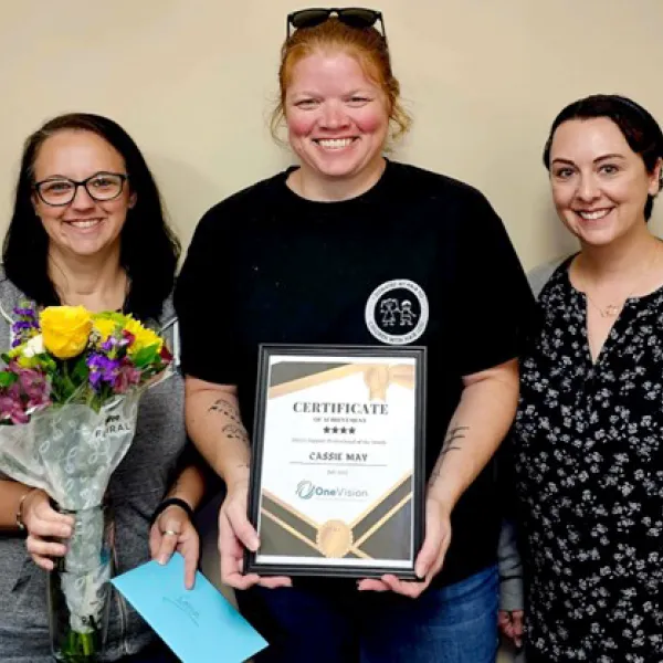 Three smiling women pose indoors with a certificate, bouquet of flowers, and an envelope, celebrating an achievement