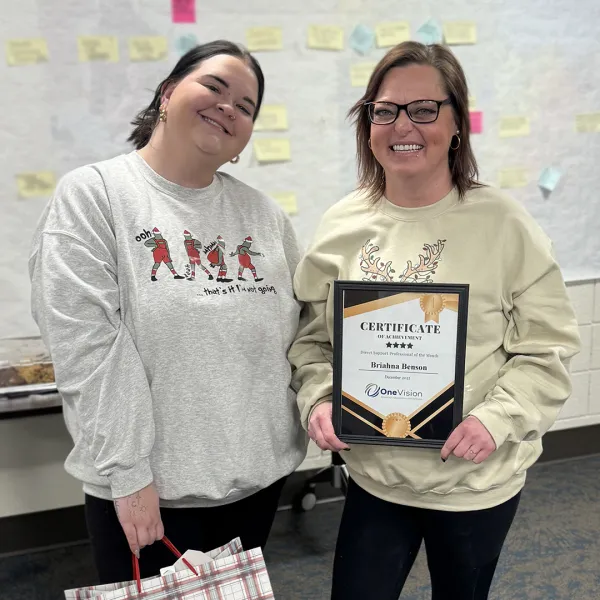 Two women smiling, one holding an achievement certificate, standing in a room with sticky notes on the wall.